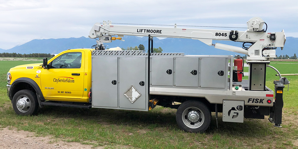 Image shows truck for hauling propane tanks with yellow cab that says CityServiceValcon, gray storage boxes and white Liftmoore crane sitting on grass with mountains in the background.