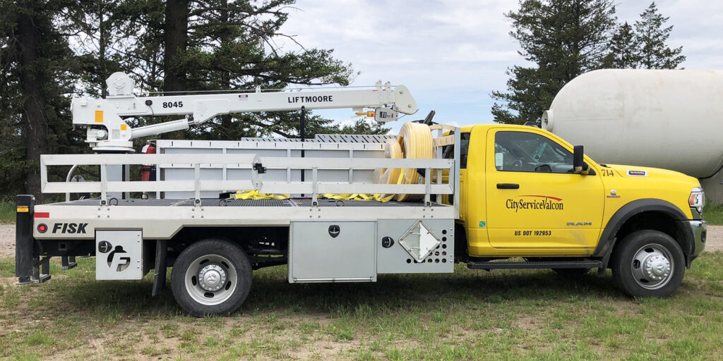 Image shows a Fisk Tank Carrier propane truck with a white liftmoore stick boom.  The truck has a yellow cab and silver metal rails on the bed, it is sitting on grass with a large propane tank and trees in the background.